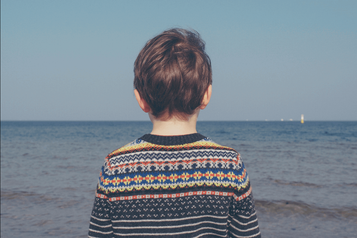 Ein Junge im Strickpulli steht am Meer und dreht uns den Rücken zu.