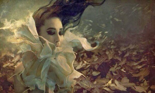 Eine Frau mit weißem Blütenkragen steht in windiger Atmosphäre auf einem Blätterteppich. 