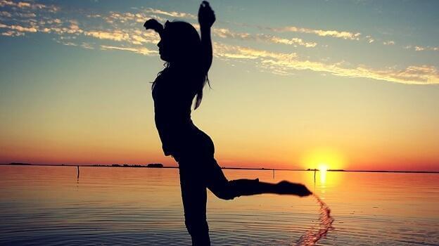 Frau im Schattenumriss tanzt im Sand vor Sonnenuntergang am Wasser.