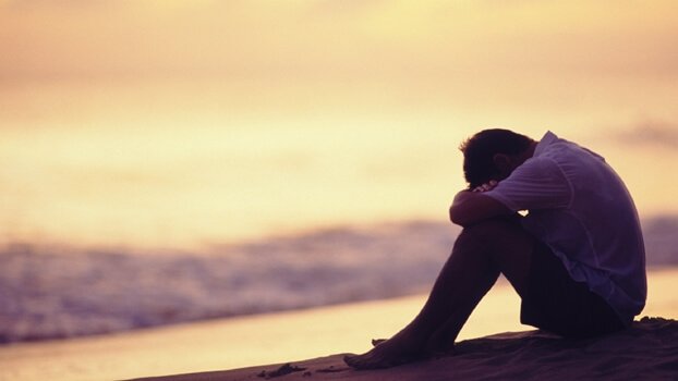 Trauriger Mann sitzt gebückt am Strand