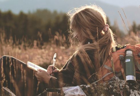 Eine Frau mit Rucksack sitzt auf einem Feld und schreibt in ein Notizbuch