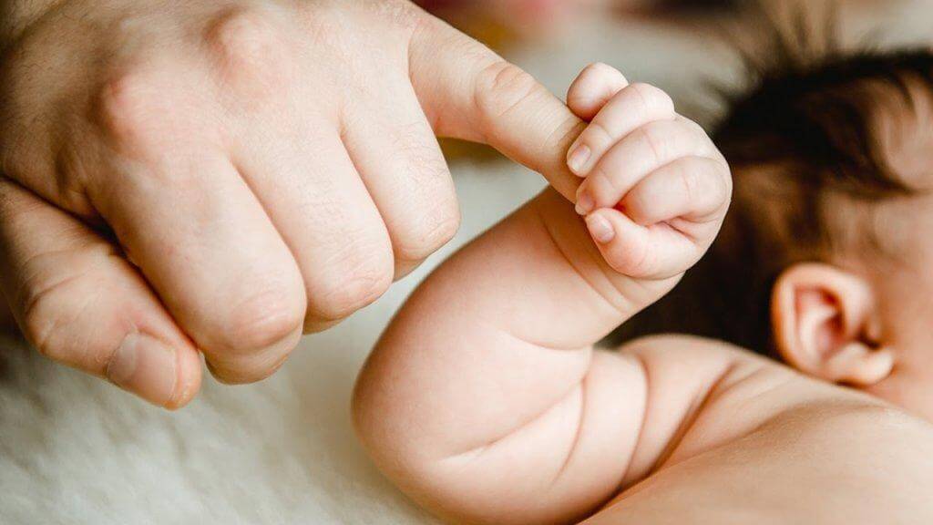 Ein Baby umfasst mit seiner rechten Hand den kleinen Finger des Vaters. 