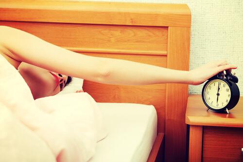 Müde Frau streckt den Arm aus dem Bett, um den Wecker auszuschalten.