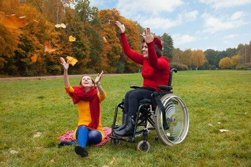 Eine junge Frau im Rollstuhl und eine Freundin amüsieren sich bei einem Ausflug ins Grüne. 