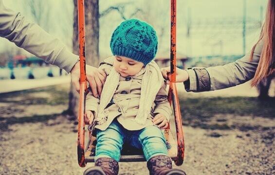 Ein kleines Mädchen sitzt auf einer Schaukel und die Eltern legen ihm jeweils eine Hand auf jede Schulter.