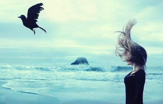 Frau steht am Meer und schaut auf einen fliegenden Raben