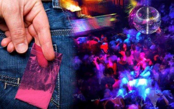 In einer Diskothek hält ein Mann, der in Jeans gekleidet ist, ein Tütchen mit einer Droge an die hintere Gesäßtasche.