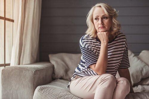 Eine Frau in der Midlife-Crisis sitzt auf einer grauen Couch, ihr Kinn auf ihrer Faust abgestützt und in die Ferne starrend.