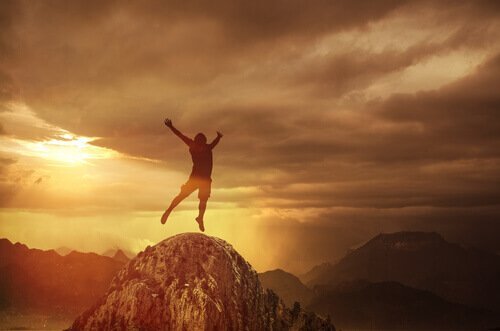Ein Mann auf einem Berggipfel, während die Sonne untergeht. Er reckt seine Arme zum Himmel. 