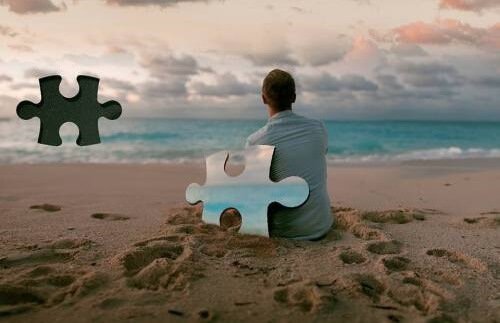 Ein Mann, angelehnt an ein Puzzelteil, sitzt am Strand und blickt auf das Meer. 