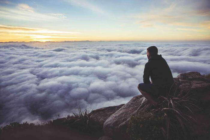 Mann sitzt auf einem Berg und schaut auf eine Wolkendecke