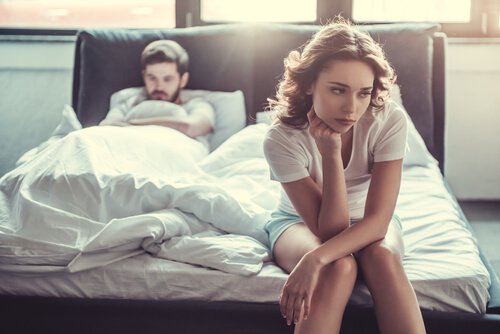Frau sitzt frustriert am Ende des Betts, ihr Partner schmollt im Hintergrund.