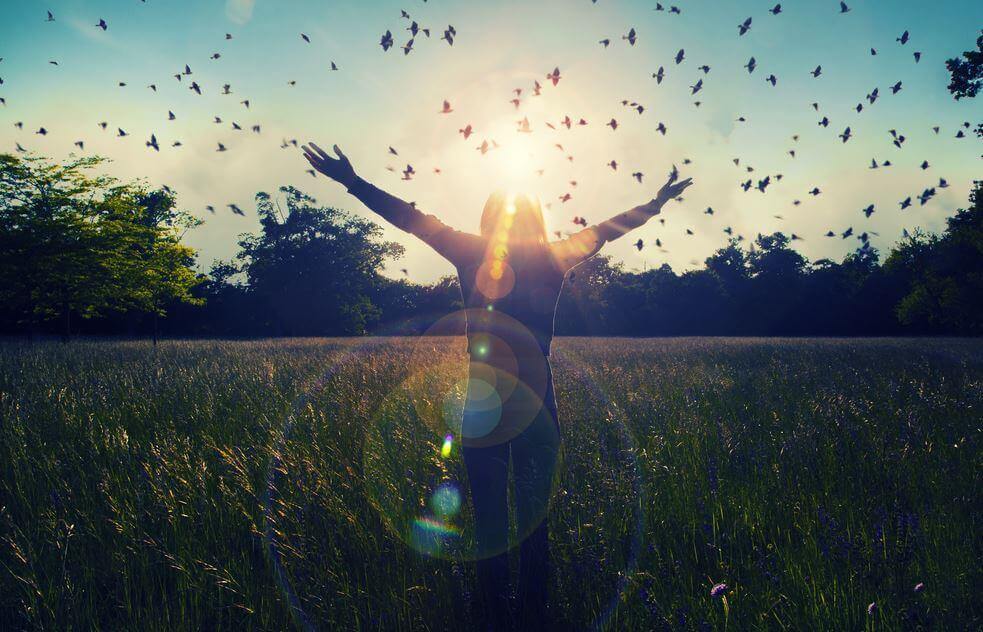 Eine Frau steht in der Natur und reckt ihre Arme in den Himmel, während ein Schwarm Vögel an ihr vorbeizieht.
