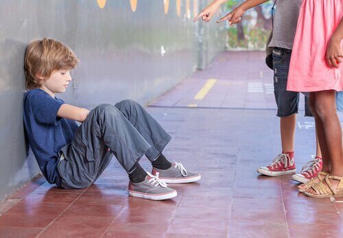 Ein trauriger Junge sitzt auf dem Fußboden und zwei Kinder stehen vor ihm und zeigen mit ihren Fingern auf den Jungen. 