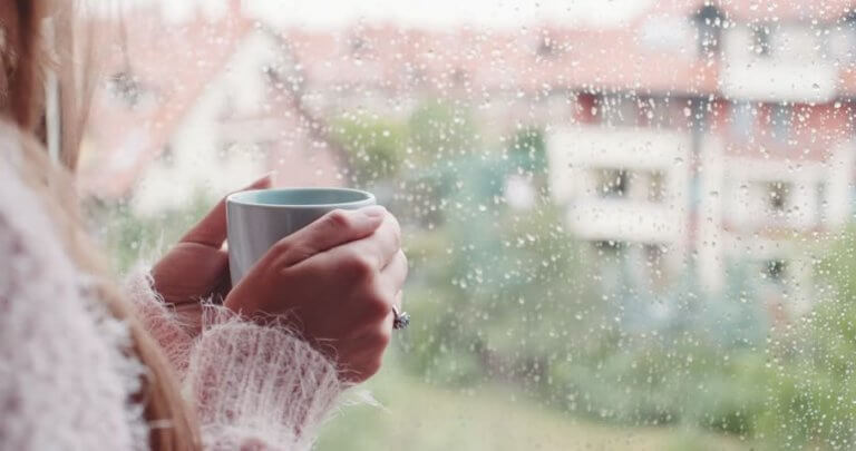 Frau trinkt Tee vor einem Fenster, auf das Regen fällt