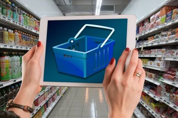 Eine Frau hält ein Tablet in einem Supermarkt hoch und der Tablet-Screen zeigt einen leeren Einkaufskorb. 