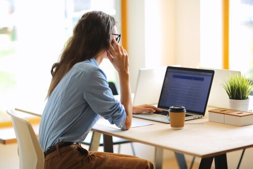 Eine Frau sitzt am Schreibtisch und telefoniert gerade, während sie auf einen Laptopscreen schaut. 