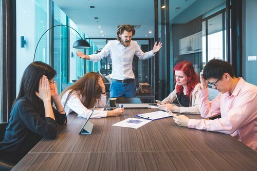 Bei einem Arbeitsmeeting ist ein Mann sichtlich ärgerlich; er ist aufgestanden und schimpft wütend.