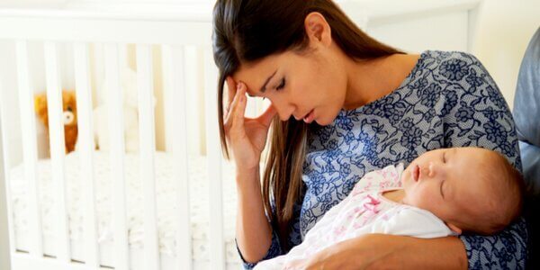 Eine junge Mutter hält ihr Baby auf dem Arm, während sie sich mit einer Hand an die Stirn greift.