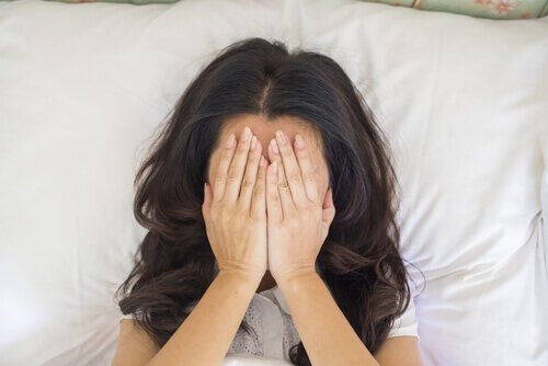 Eine Frau liegt gestresst im Bett und hat die Hände vor ihr Gesicht geschlagen.
