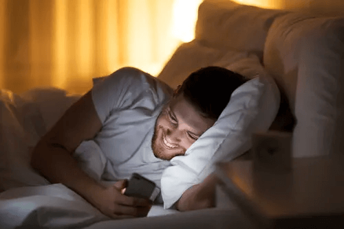 Mann liegt lachend mit Handy im Bett